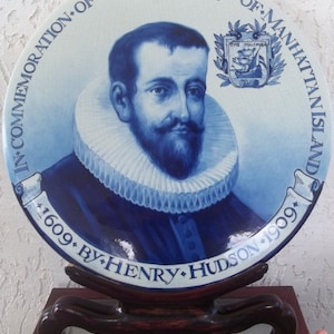 May include: A blue and white commemorative plate featuring a portrait of Henry Hudson. The plate includes text that reads "In Commemoration of the Discovery of Manhattan Island 1609 by Henry Hudson 1909." The plate is displayed on a wooden stand.
