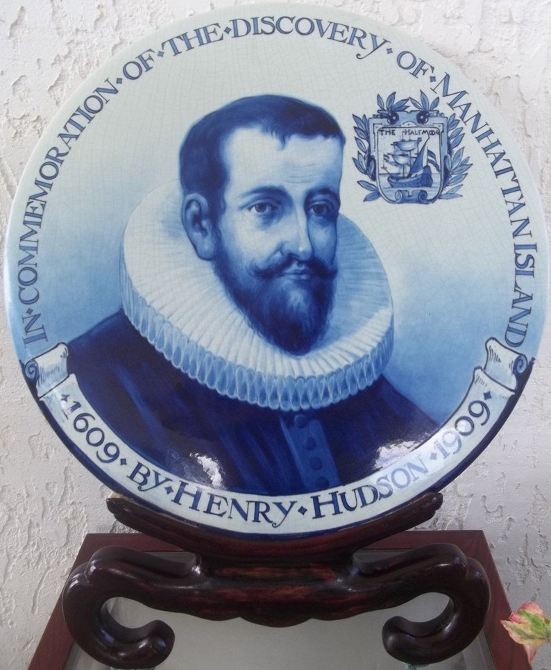May include: A blue and white commemorative plate featuring a portrait of Henry Hudson. The plate includes text that reads "In Commemoration of the Discovery of Manhattan Island 1609 by Henry Hudson 1909." The plate is displayed on a wooden stand.