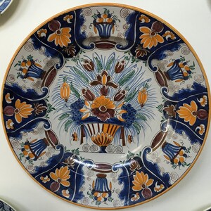 Puede incluir: Un plato grande de cer&aacute;mica blanca con un dise&ntilde;o floral azul y naranja. El plato presenta un motivo floral central con un jarr&oacute;n y un borde de flores y hojas estilizadas.