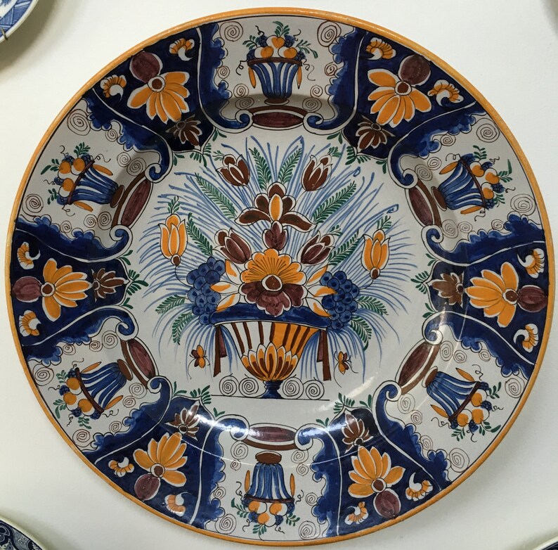 Puede incluir: Un plato grande de cer&aacute;mica blanca con un dise&ntilde;o floral azul y naranja. El plato presenta un motivo floral central con un jarr&oacute;n y un borde de flores y hojas estilizadas.