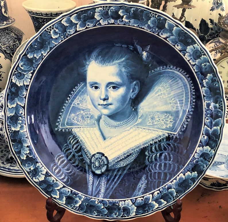 Pu&ograve; includere: Un piatto in ceramica blu e bianco con un ritratto di una giovane donna che indossa un colletto di pizzo. Il piatto &egrave; decorato con motivi floreali intorno al bordo.