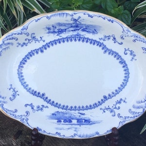 May include: An oval ceramic serving platter featuring a white base with blue transferware patterns. The design includes a central oval border, intricate scrollwork, and landscape scenes with windmills. The rim is finished with a thin gold trim.