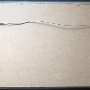May include: The back of a framed picture with a wire hanging system. The frame is black and the backing is a light brown cardboard.