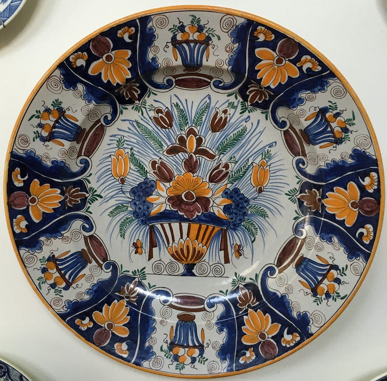 May include: A decorative ceramic plate with a floral design. The plate features a white background with a central arrangement of flowers in shades of orange, brown, and blue. The rim is bordered with orange and blue floral patterns.