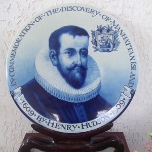 Puede incluir: Plato conmemorativo azul y blanco con un retrato de Henry Hudson. El plato est&aacute; inscrito con texto, incluyendo "In Commemoration of the Discovery of Manhattan Island 1609 by Henry Hudson 1909". El plato se exhibe en un soporte de madera oscura.