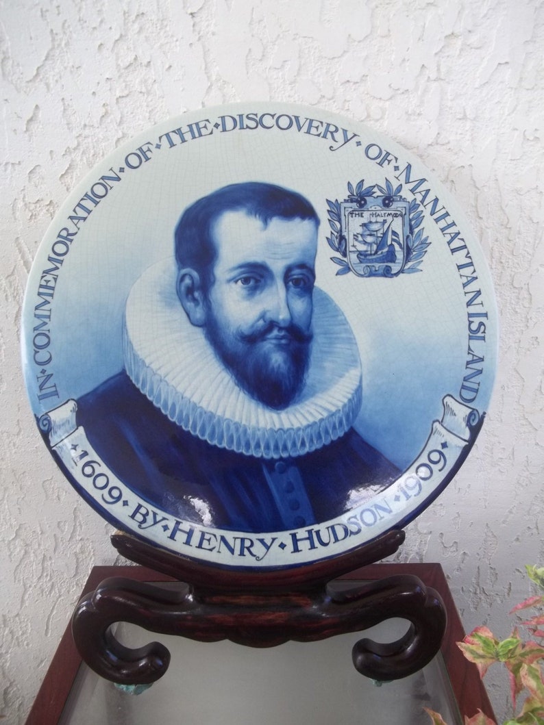 Puede incluir: Plato conmemorativo azul y blanco con un retrato de Henry Hudson. El plato est&aacute; inscrito con texto, incluyendo "In Commemoration of the Discovery of Manhattan Island 1609 by Henry Hudson 1909". El plato se exhibe en un soporte de madera oscura.
