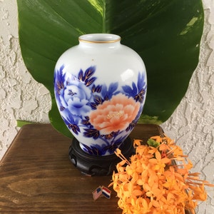 May include: A white porcelain vase with a gold rim, decorated with blue and peach floral patterns. The vase is on a dark wooden stand. A small bunch of orange flowers and two flag pins are beside it.