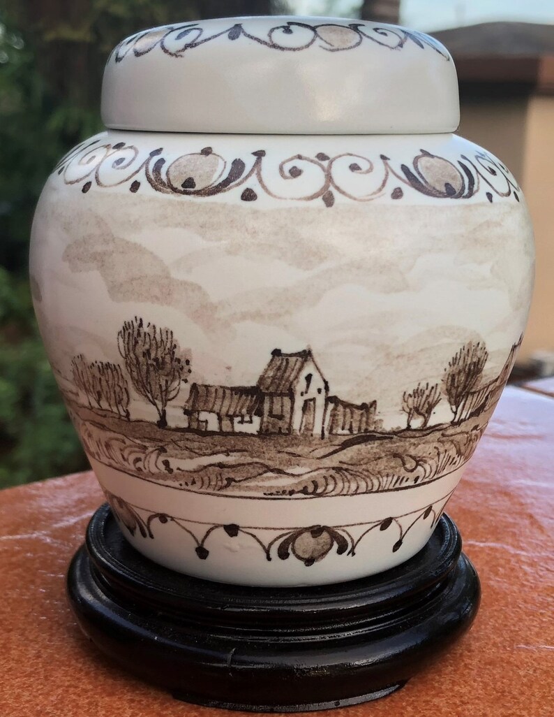 May include: A white ceramic jar with a lid, featuring a hand-painted landscape scene in brown tones. The jar sits on a black wooden stand. Decorative brown scrollwork accents the top and bottom rims. The jar has a round shape.