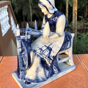K&ouml;nnte beinhalten: Eine wei&szlig;-blaue Keramikfigur, die eine Frau an einem Spinnrad darstellt. Die Frau tr&auml;gt eine Haube und eine Sch&uuml;rze mit einer Windm&uuml;hlen-Szene. Die Figur steht auf einem rechteckigen Sockel. Detailliertes Design.
