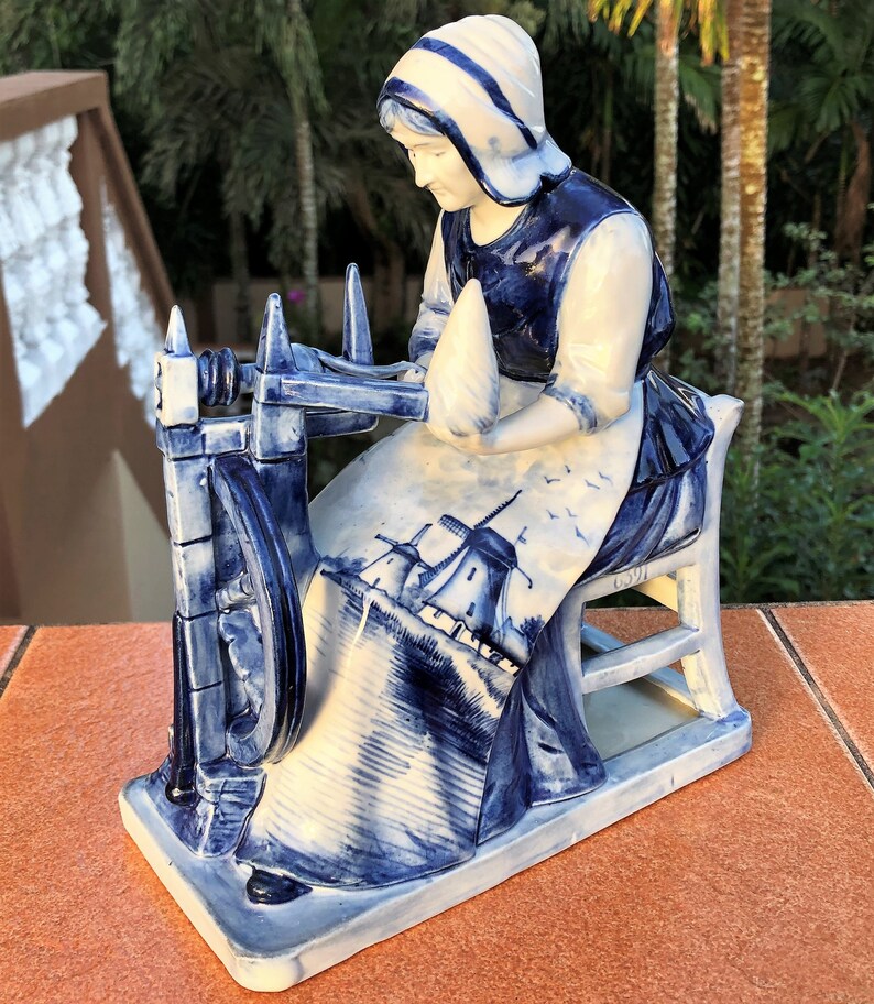K&ouml;nnte beinhalten: Eine wei&szlig;-blaue Keramikfigur, die eine Frau an einem Spinnrad darstellt. Die Frau tr&auml;gt eine Haube und eine Sch&uuml;rze mit einer Windm&uuml;hlen-Szene. Die Figur steht auf einem rechteckigen Sockel. Detailliertes Design.