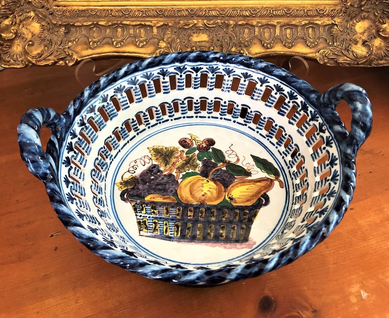 Może przedstawiać: Ręcznie malowana ceramiczna misa na owoce z niebiesko-białym wzorem. Misa ma pośrodku kosz owoc&oacute;w, w tym winogrona, gruszki i wiśnie. Obręcz ma dekoracyjny wz&oacute;r i dwa uchwyty.