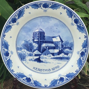May include: A white ceramic plate with a blue and white church scene. The plate has a blue floral border and the text "KERSTMIS 1970". The plate is displayed on a wooden stand.