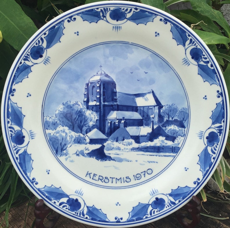 May include: A white ceramic plate with a blue and white church scene. The plate has a blue floral border and the text "KERSTMIS 1970". The plate is displayed on a wooden stand.