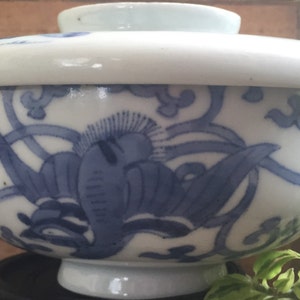 May include: A white and blue ceramic bowl with a lid. The bowl features a blue floral design and a white rim. The bowl is likely used for serving food or as a decorative item. The bowl is sitting on a black stand.