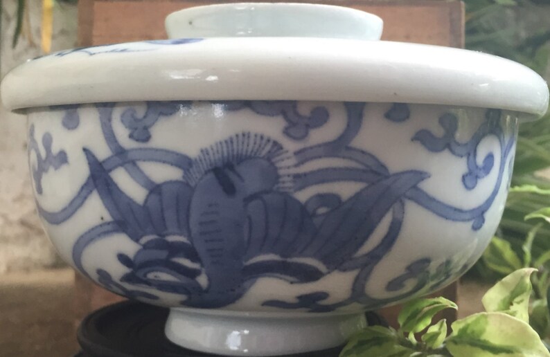 May include: A white and blue ceramic bowl with a lid. The bowl features a blue floral design and a white rim. The bowl is likely used for serving food or as a decorative item. The bowl is sitting on a black stand.