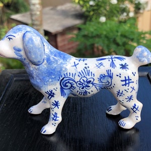 Puede incluir: Figura de perro de cer&aacute;mica en posici&oacute;n de pie, con base blanca y dise&ntilde;os florales y abstractos azules. Las orejas y partes del cuerpo del perro est&aacute;n pintadas de azul. La figura tiene un acabado brillante.