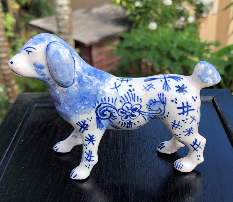 Puede incluir: Figura de perro de cer&aacute;mica en posici&oacute;n de pie, con base blanca y dise&ntilde;os florales y abstractos azules. Las orejas y partes del cuerpo del perro est&aacute;n pintadas de azul. La figura tiene un acabado brillante.