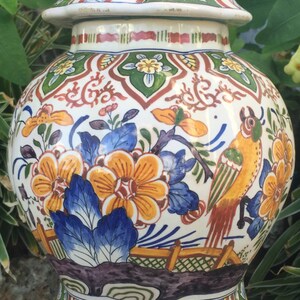 May include: A decorative ceramic jar with a lid, featuring a vibrant floral and bird design. The jar is predominantly white with orange, blue, green, and red accents. It sits on a dark wooden base, showcasing a detailed, hand-painted aesthetic.
