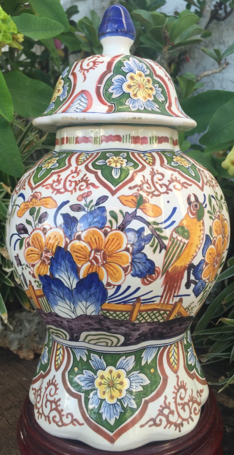 May include: A decorative ceramic jar with a lid, featuring a vibrant floral and bird design. The jar is predominantly white with orange, blue, green, and red accents. It sits on a dark wooden base, showcasing a detailed, hand-painted aesthetic.