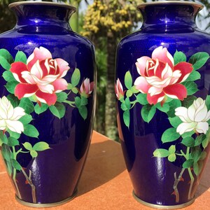 May include: Two cobalt blue vases with floral designs. Each vase features a large red and pink rose, a white flower, and green leaves. The vases have a metallic rim at the top and base, showcasing an elegant aesthetic.