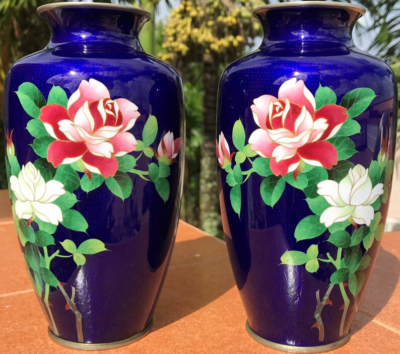 May include: Two cobalt blue vases with floral designs. Each vase features a large red and pink rose, a white flower, and green leaves. The vases have a metallic rim at the top and base, showcasing an elegant aesthetic.