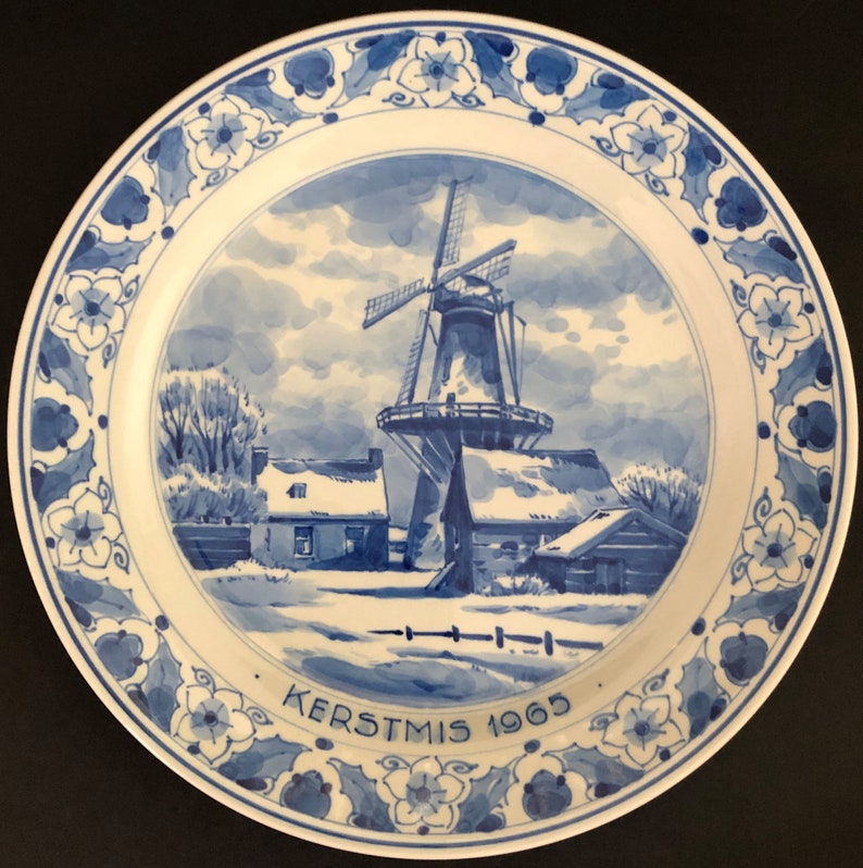 May include: Blue and white Delftware plate featuring a winter scene of a windmill and houses. The plate has a blue and white floral border and the text "KERSTMIS 1965".