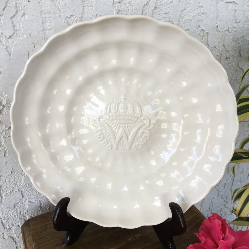Puede incluir: Plato de cer&aacute;mica blanco con un dise&ntilde;o en relieve de una corona y la letra "W" en el centro. El plato tiene un borde festoneado.