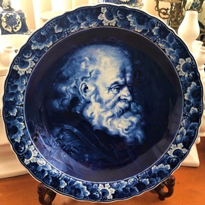 May include: A blue and white ceramic plate featuring a detailed portrait of a bearded man. The plate has a scalloped edge and is decorated with floral designs.