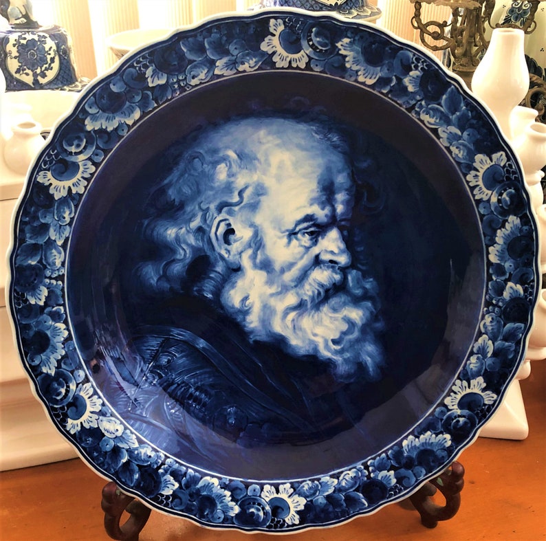 May include: A blue and white ceramic plate featuring a detailed portrait of a bearded man. The plate has a scalloped edge and is decorated with floral designs.