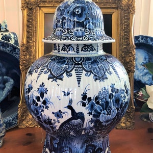 May include: A large blue and white Delftware vase with a peacock design. The vase has a round body and a tall, domed lid. The lid is decorated with floral motifs. The vase is sitting on a wooden table.