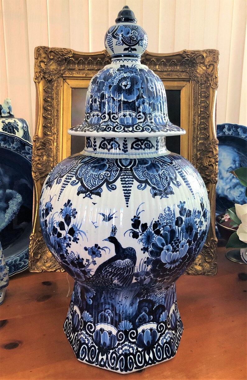 May include: A large blue and white Delftware vase with a peacock design. The vase has a round body and a tall, domed lid. The lid is decorated with floral motifs. The vase is sitting on a wooden table.