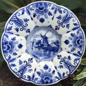 May include: A white ceramic plate with a blue and white windmill scene in the center. The plate has a scalloped edge and is decorated with blue and white floral designs.