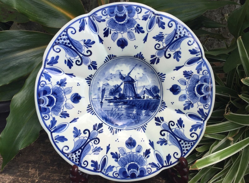 May include: A white ceramic plate with a blue and white windmill scene in the center. The plate has a scalloped edge and is decorated with blue and white floral designs.