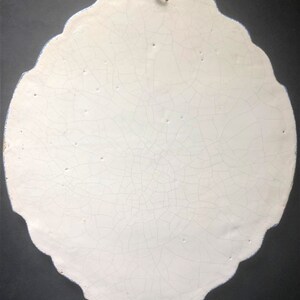 May include: A white, oval-shaped ceramic wall hanging with a scalloped edge and a blue border. The surface is heavily crackled, and a small metal hook is attached at the top for hanging. The overall aesthetic is vintage.