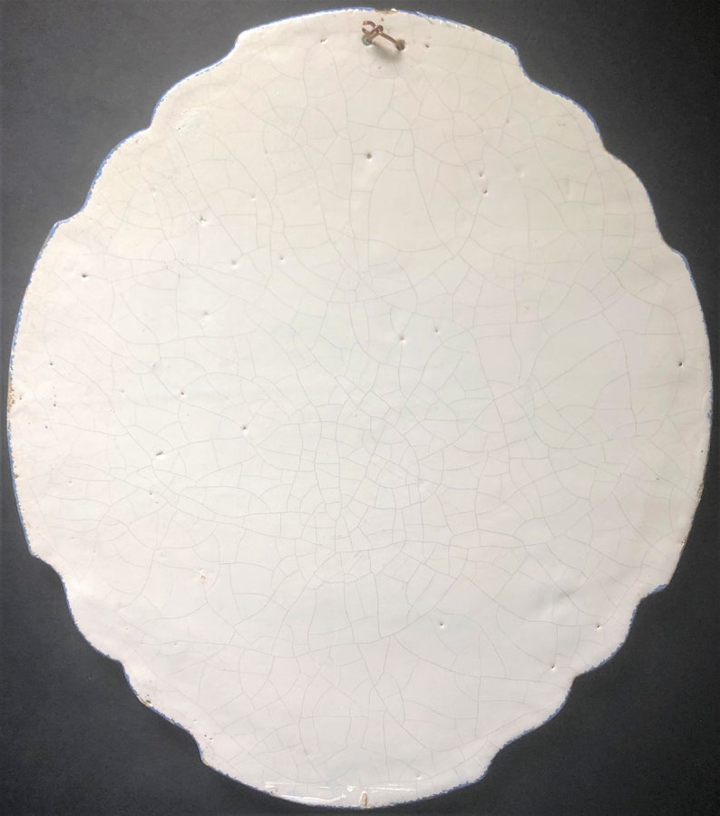 May include: A white, oval-shaped ceramic wall hanging with a scalloped edge and a blue border. The surface is heavily crackled, and a small metal hook is attached at the top for hanging. The overall aesthetic is vintage.