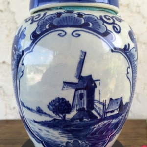 May include: A blue and white ceramic ginger jar with a lid. The jar features a windmill scene in blue, surrounded by floral designs. The lid and base have blue accents. The jar is a decorative item.