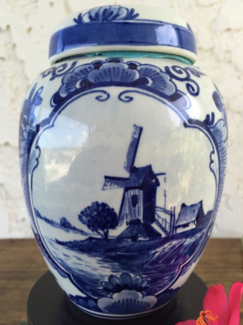 May include: A blue and white ceramic ginger jar with a lid. The jar features a windmill scene in blue, surrounded by floral designs. The lid and base have blue accents. The jar is a decorative item.