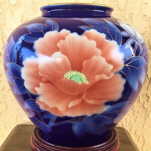 May include: A cobalt blue ceramic vase with a floral design. The vase features a large, pink peony with gold accents and green leaves. The vase is sitting on a wooden base.