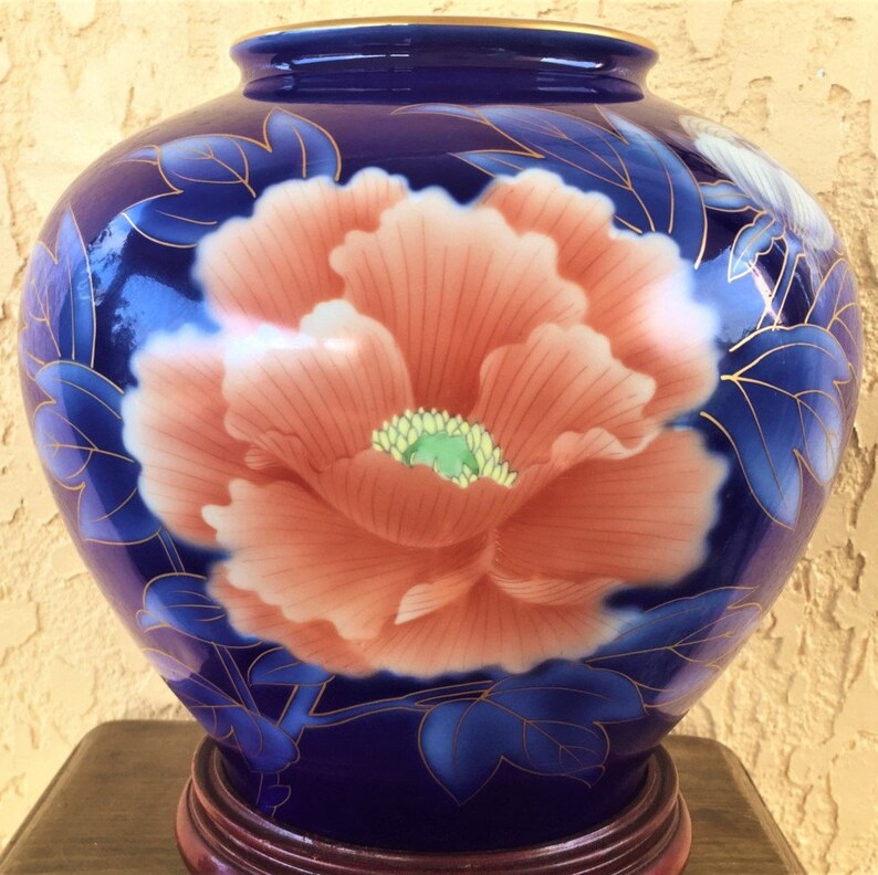 May include: A cobalt blue ceramic vase with a floral design. The vase features a large, pink peony with gold accents and green leaves. The vase is sitting on a wooden base.