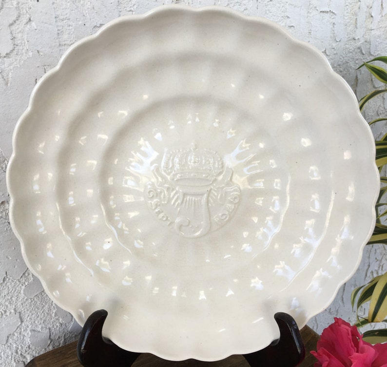 Puede incluir: Un plato de cer&aacute;mica blanco con un borde festoneado y un dise&ntilde;o en relieve en el centro. El dise&ntilde;o presenta una corona, un s&iacute;mbolo y la palabra "OSPEDALE".