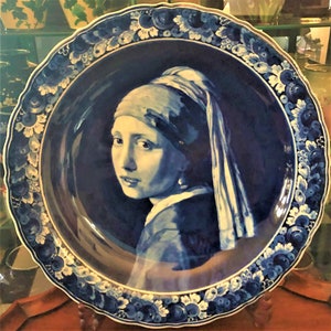 May include: A decorative blue and white ceramic plate featuring a portrait of a woman with a pearl earring. The plate has a scalloped edge with floral patterns and a dark blue background.