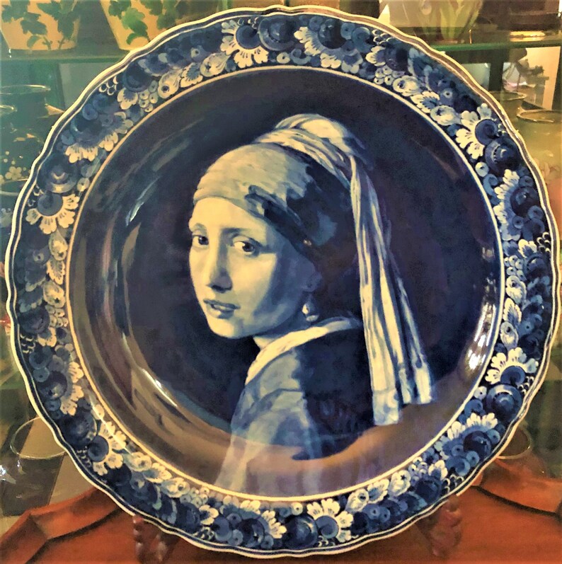May include: A decorative blue and white ceramic plate featuring a portrait of a woman with a pearl earring. The plate has a scalloped edge with floral patterns and a dark blue background.