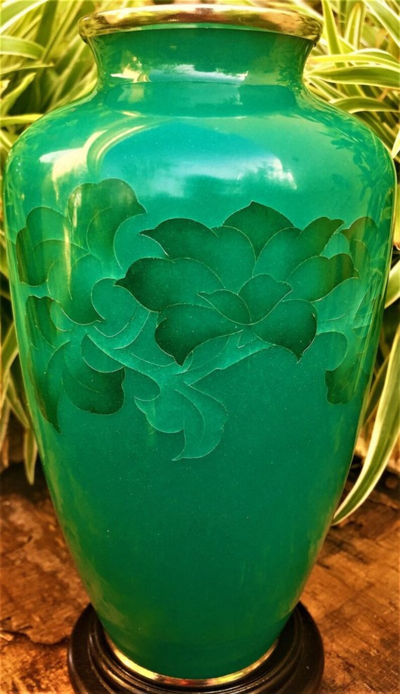 May include: A green vase with a floral pattern and a gold rim. The vase has a glossy finish. The floral design is a darker shade of green. The vase is set on a dark brown base. The vase is likely made of ceramic or glass.