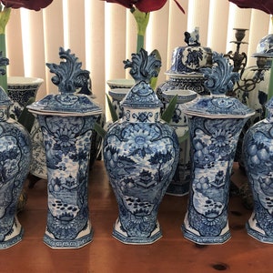 May include: A collection of blue and white ceramic vases with lids. The vases feature a detailed landscape design with floral and architectural elements. The lids are topped with a decorative finial. The vases are arranged on a wooden surface.