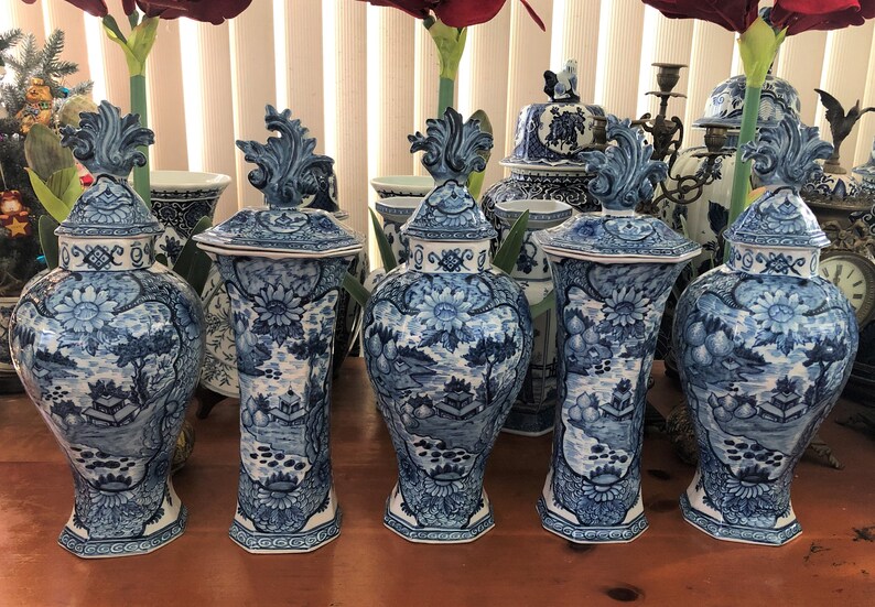 May include: A collection of blue and white ceramic vases with lids. The vases feature a detailed landscape design with floral and architectural elements. The lids are topped with a decorative finial. The vases are arranged on a wooden surface.