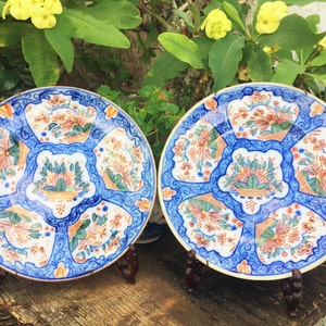 K&ouml;nnte beinhalten: Zwei antike blaue und wei&szlig;e Keramikteller mit floralen Mustern in Orange, Gr&uuml;n und Gold. Die Teller stehen auf Holzst&auml;ndern.