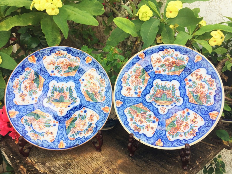 K&ouml;nnte beinhalten: Zwei antike blaue und wei&szlig;e Keramikteller mit floralen Mustern in Orange, Gr&uuml;n und Gold. Die Teller stehen auf Holzst&auml;ndern.