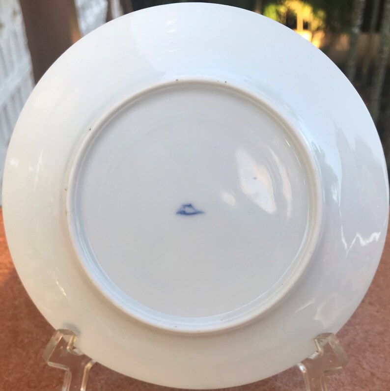 May include: A white porcelain plate with a blue emblem on the underside. The plate has a circular design with a slightly raised edge. The plate is displayed on a clear stand.