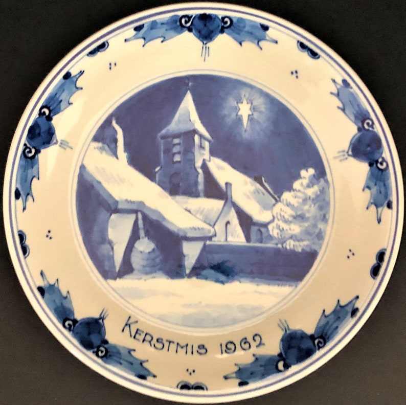 May include: A blue and white ceramic plate featuring a winter scene of a church and houses covered in snow. The plate has a blue and white border with a holly leaf design. The text "KERSTMIS 1962" is written on the bottom of the plate.