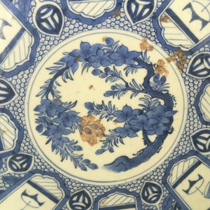 Op de afbeelding: Een blauw en wit keramisch bord met een bloemenontwerp en geometrische patronen. Het bord heeft een centraal ontwerp van een boom met bloemen en bladeren, omringd door een rand van zich herhalende geometrische vormen.